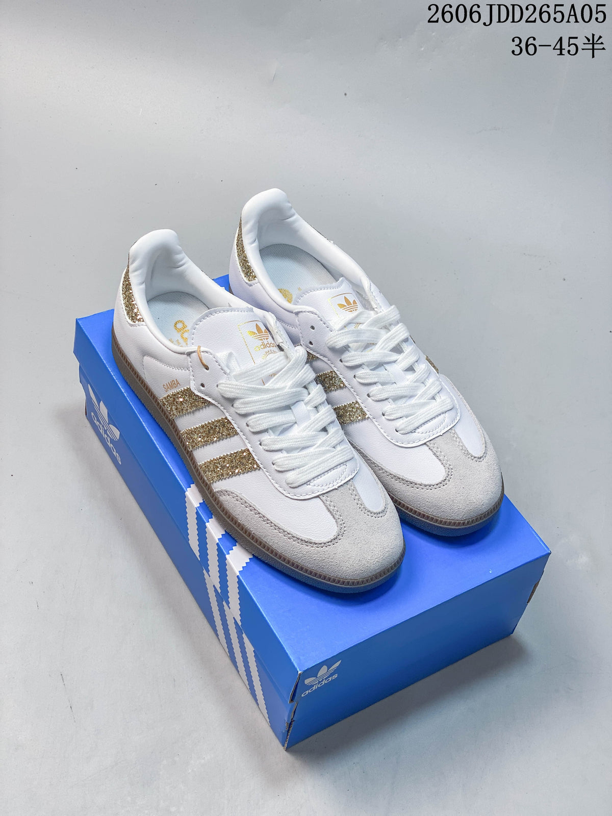 Pair of white adidas Samba OG low-top sneakers with gold glitter stripes and gum-colored soles on a blue shoebox.