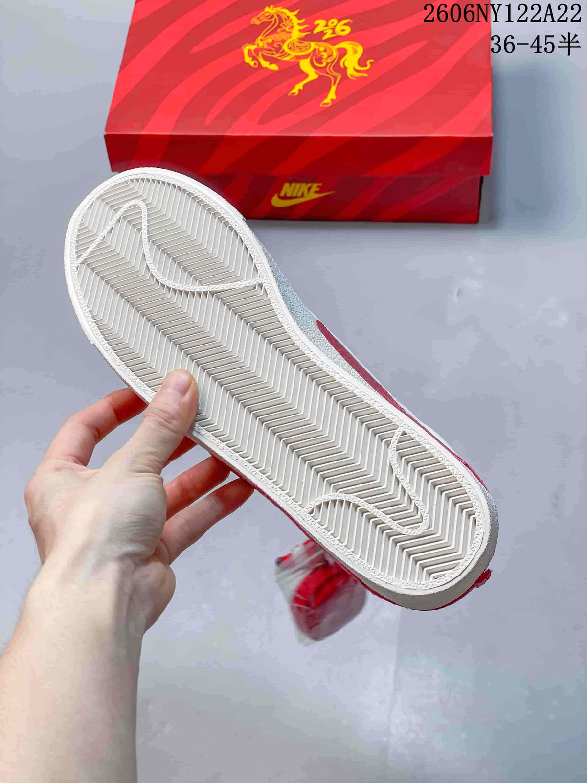 Underside of the white sneaker showing a herringbone-pattern outsole, with the red shoebox behind it.