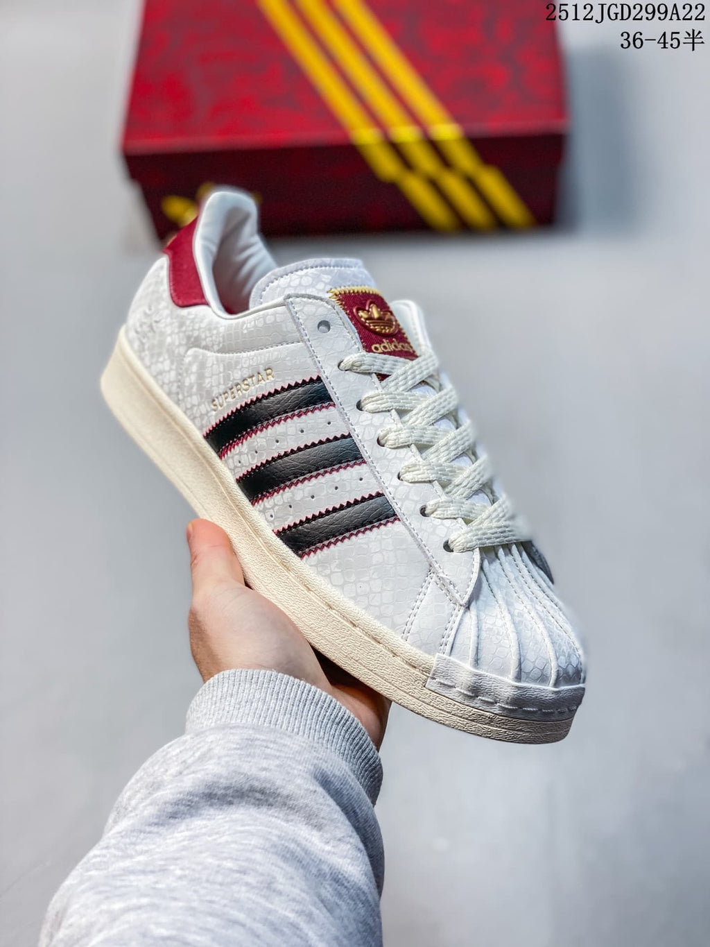 Angled view of a white patterned Superstar sneaker with black side stripes and burgundy details.