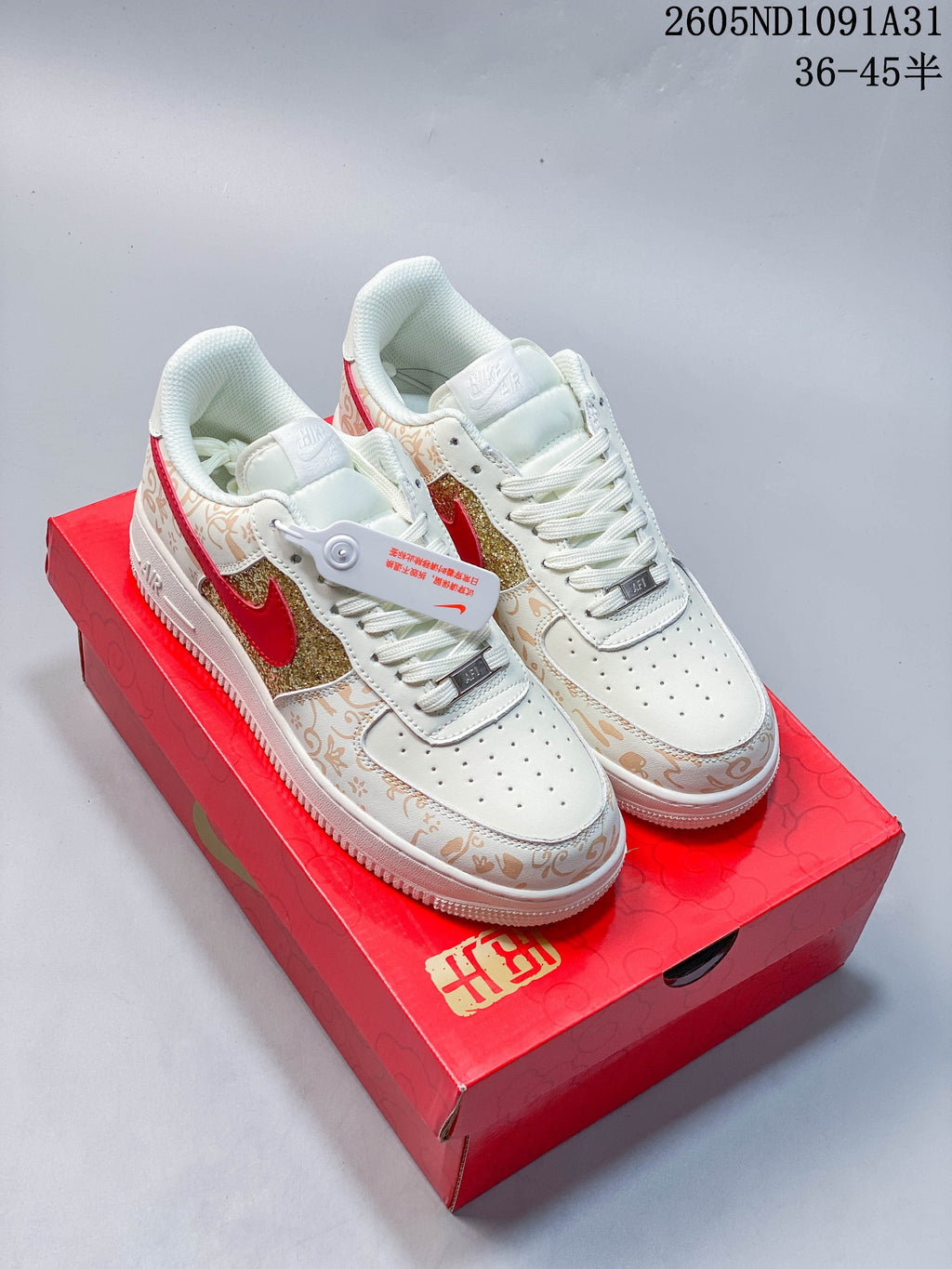 Pair of white low-top sneakers with red Swooshes, gold glitter panels and tan decorative patterns, shown on a red shoe box.