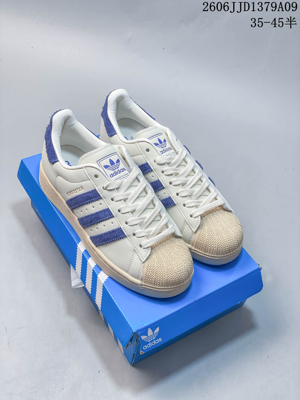 Pair of cream adidas Superstar sneakers with blue accents and shell toes on a blue shoe box.