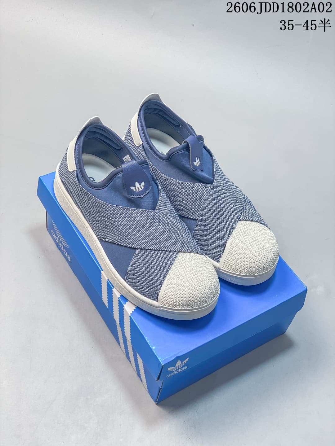 Pair of blue and white Adidas slip-on sneakers displayed on a blue Adidas shoebox, showing the shell toes and Trefoil branding.