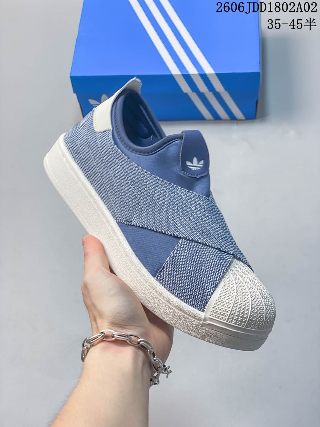 Blue and white Adidas slip-on sneaker shown from the side with a textured shell toe, crossover upper panels, and a blue shoebox behind it.