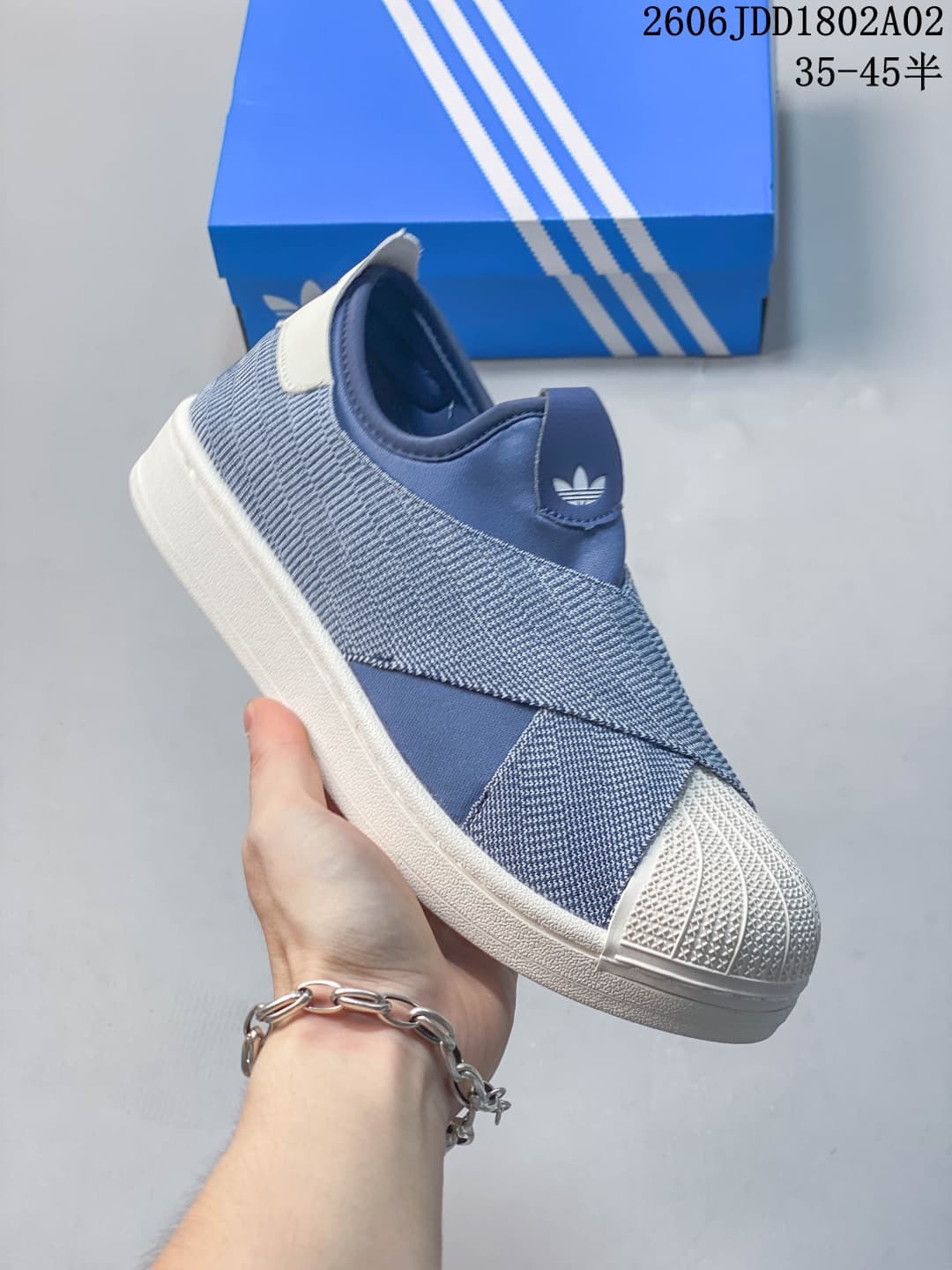 Blue and white Adidas slip-on sneaker shown from the side with a textured shell toe, crossover upper panels, and a blue shoebox behind it.