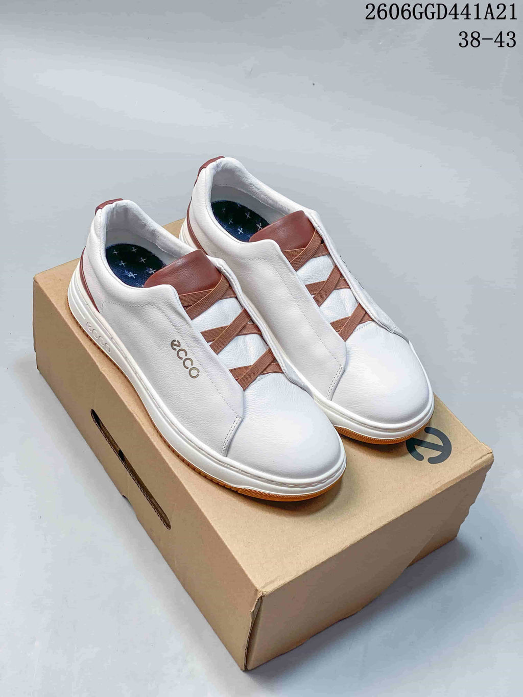 White ECCO low-top slip-on sneakers with brown elastic straps, shown on a box.