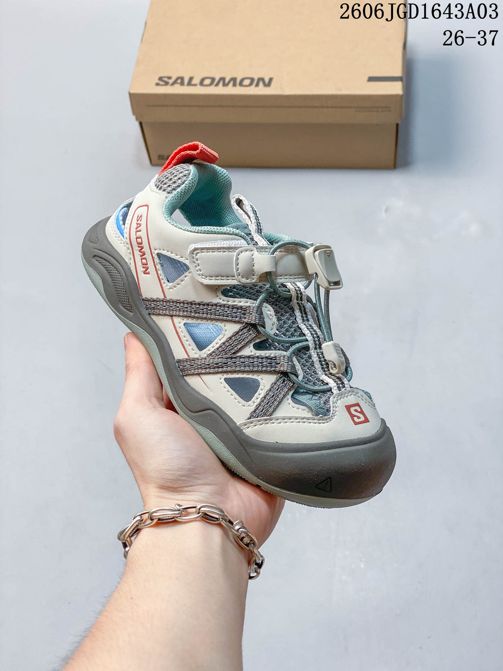 Side view of a light gray and blue-gray kids’ Salomon sneaker with cord lacing and a hook-and-loop strap.