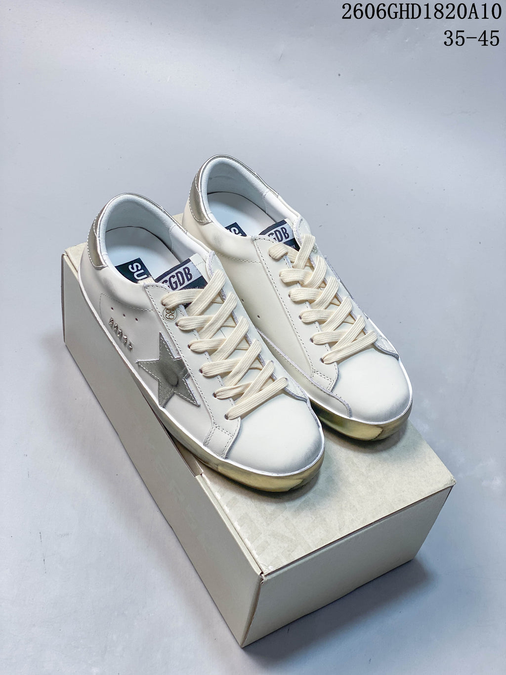 Pair of white Golden Goose Super-Star style low-top sneakers with silver star appliqués and metallic heel details, shown on a box.