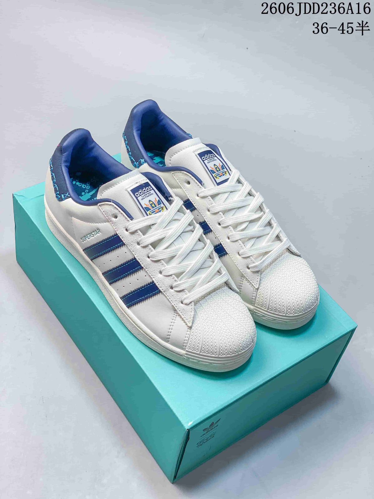 White Adidas Originals Superstar sneakers with blue stripes and shell toes shown on a turquoise box.