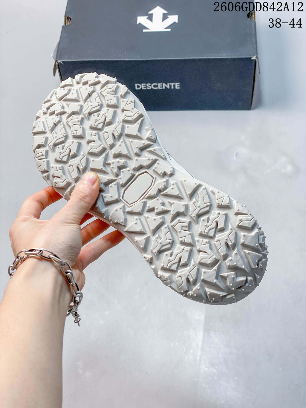 Close-up of the white sneaker's deeply patterned outsole beside a DESCENTE shoe box; the image displays size range 38–44.