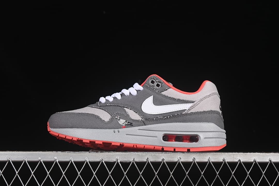 Side view of grey Nike Air Max 1 sneakers with a white Swoosh, crimson accents, and a visible heel Air unit.