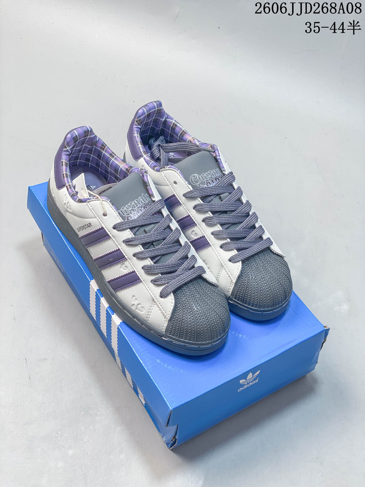 White and purple Superstar 2 sneakers with plaid lining and dark shell toes, pictured on a blue shoe box.