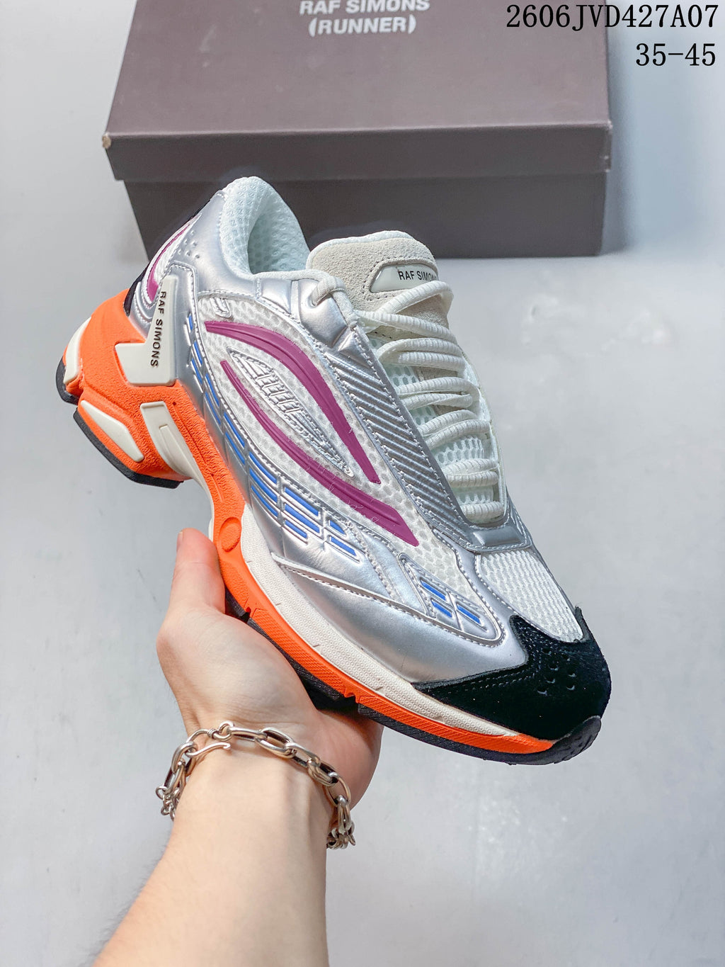 Angled side view of a silver and white sneaker with magenta accents, dark toe panel, and bright orange outsole; image overlay shows 35–45.