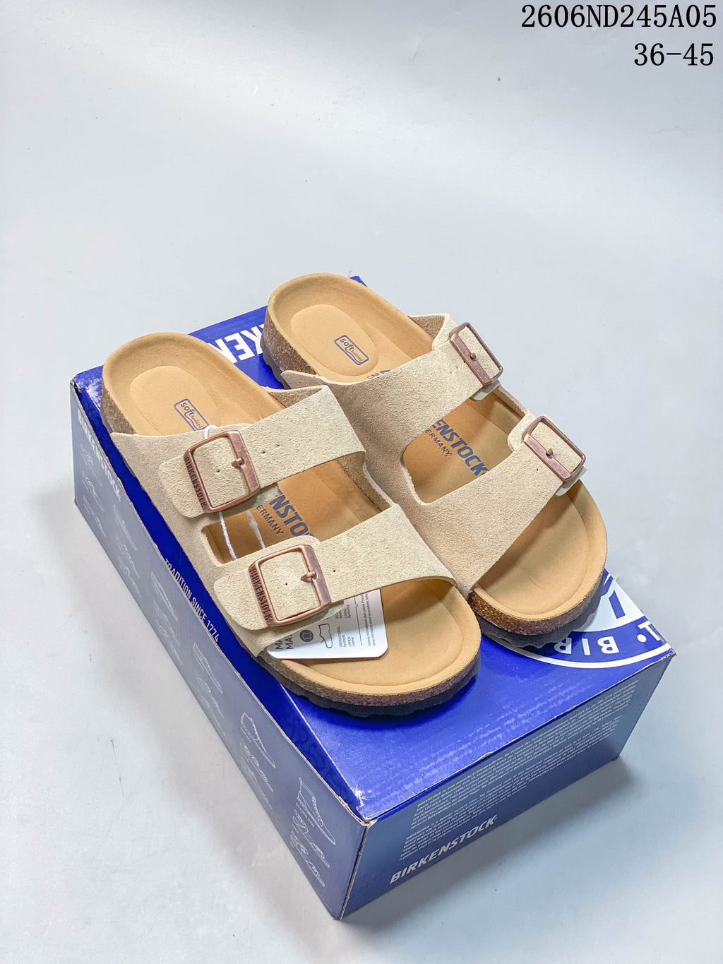 Pair of beige two-strap sandals with adjustable buckles shown on a blue shoebox.