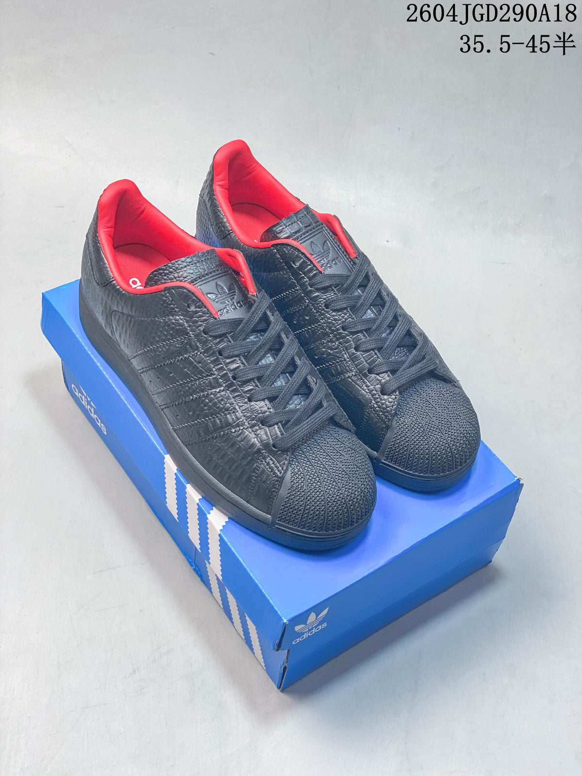 Pair of black textured adidas Originals Superstar 2 sneakers with red lining, shown on a blue shoe box.