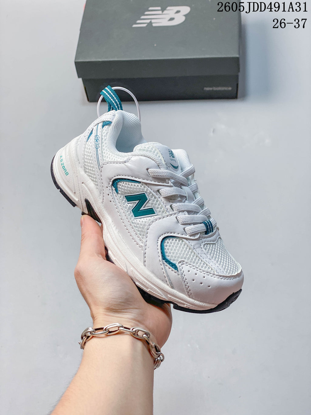 White kids' New Balance 530 lace-up sneaker with teal accents, shown in hand.