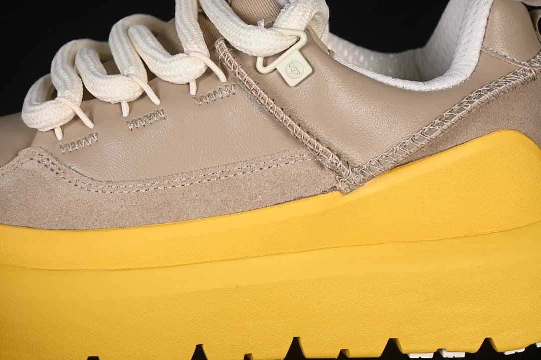 Close-up of taupe upper panels, stitched seams, cream laces, and yellow sole