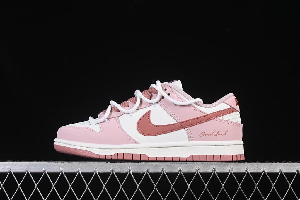 Side view of a pink and white Nike SB Dunk Low sneaker with dusty-rose accents and “Good Luck” lettering on the heel.