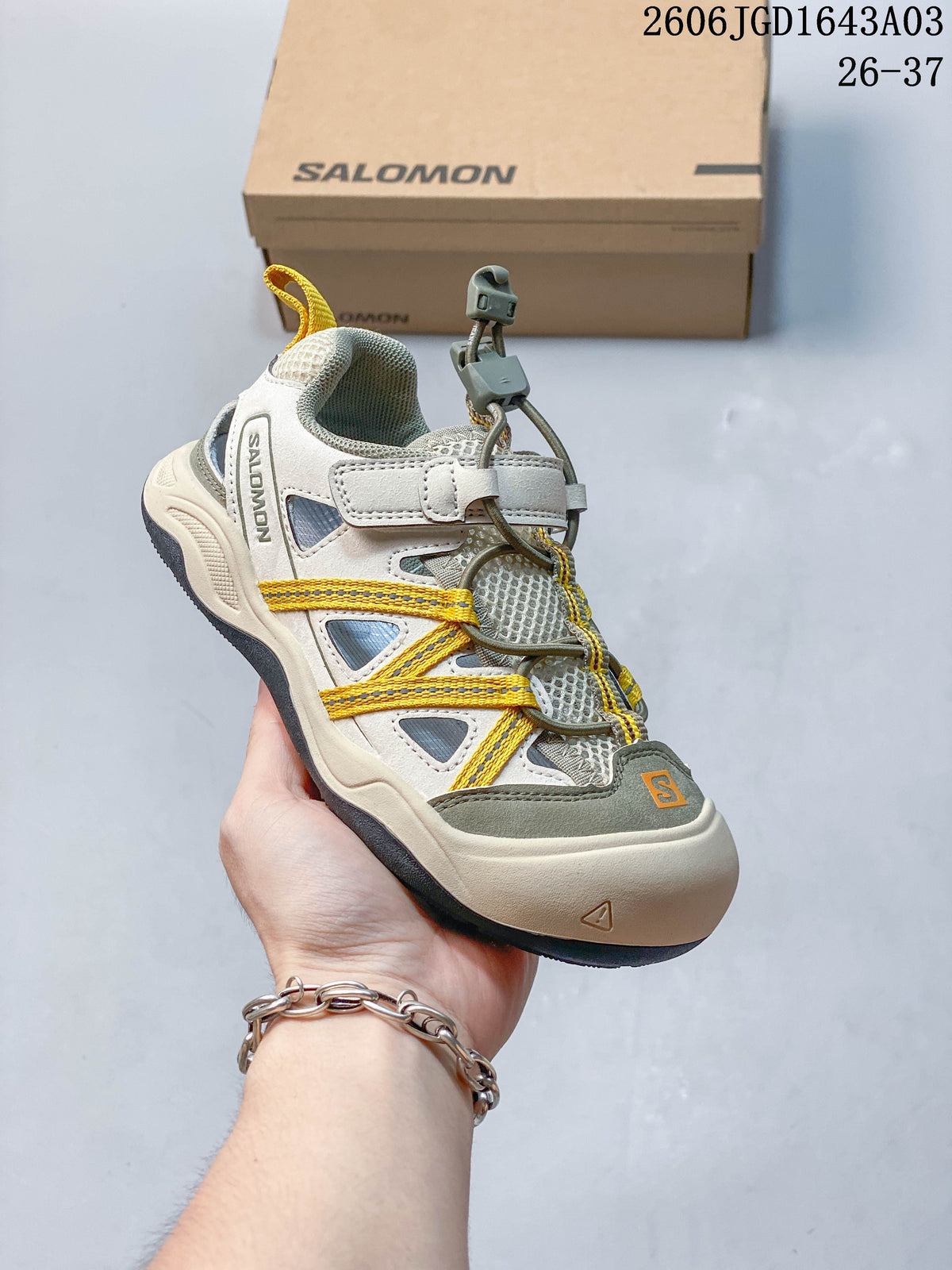 Side view of a light neutral kids’ Salomon sneaker with yellow cord accents and a toggle closure.