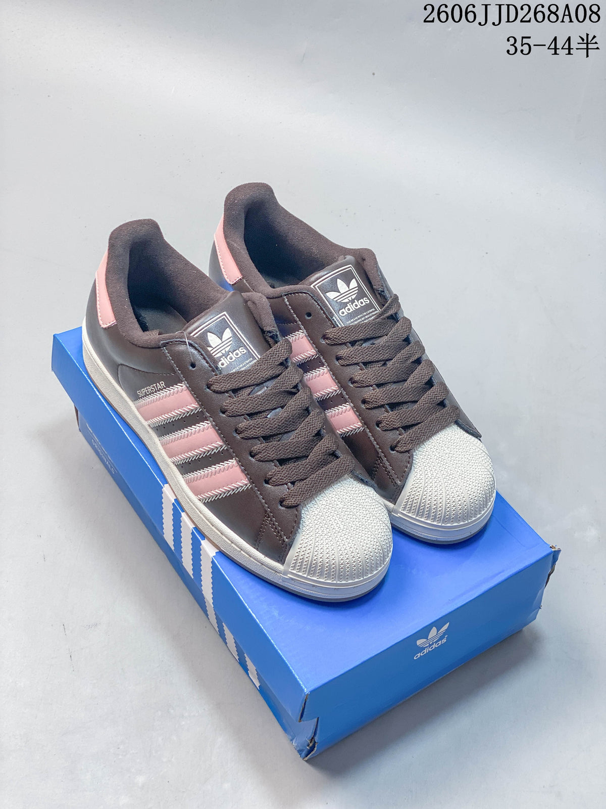 Dark brown adidas Superstar 2 sneakers with pink stripes and light shell toes, pictured on a blue shoe box.