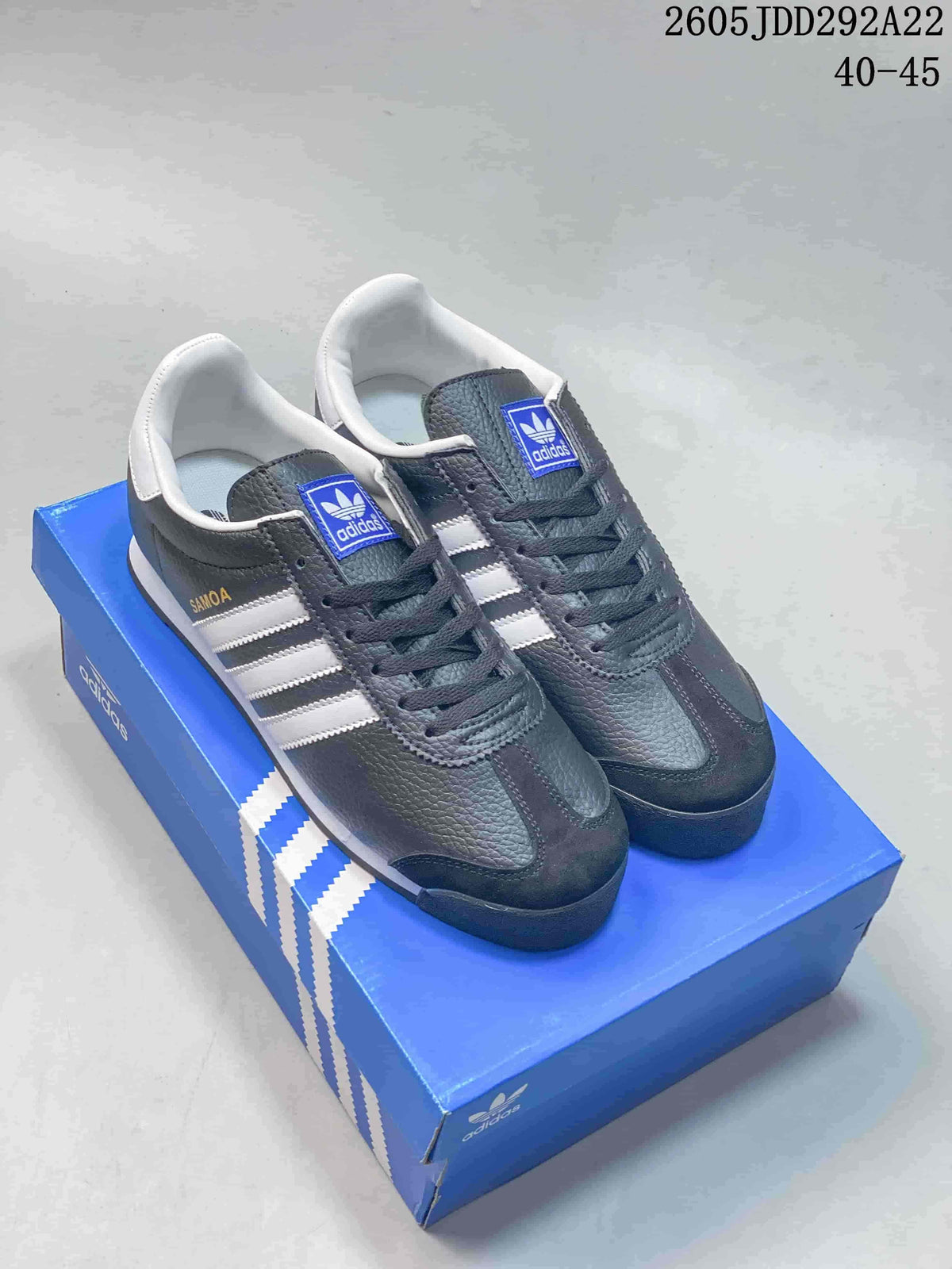 Pair of charcoal Adidas Samoa low-top sneakers with white stripes and dark laces on a blue shoe box