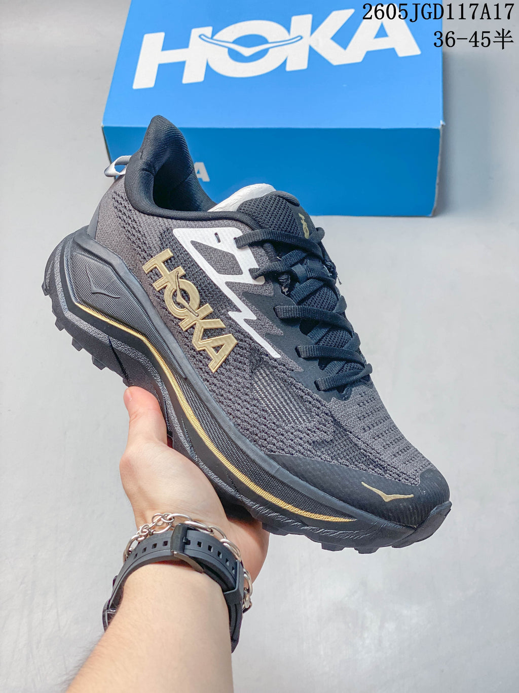 Dark gray Hoka Mafate Speed 2 shoe with gold accents, shown with a blue shoe box.
