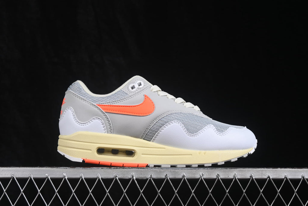 Side profile of white and grey Nike Air Max 1 sneakers with orange Swoosh branding and a visible Air unit.