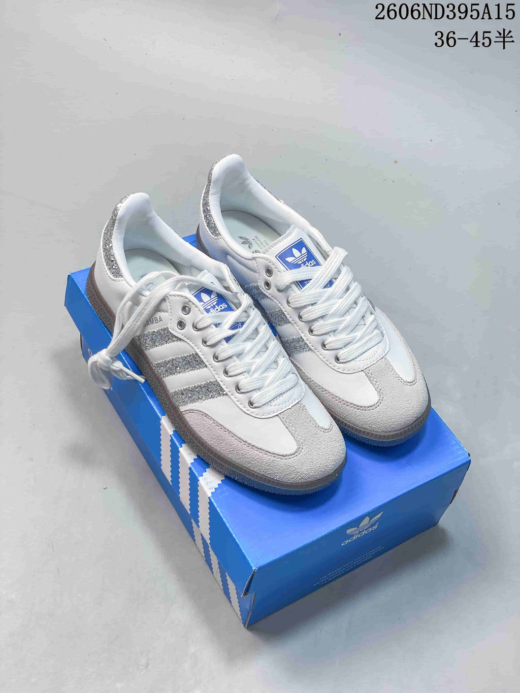 Pair of white Adidas Samba low-top sneakers with silver glitter accents displayed on a blue shoebox