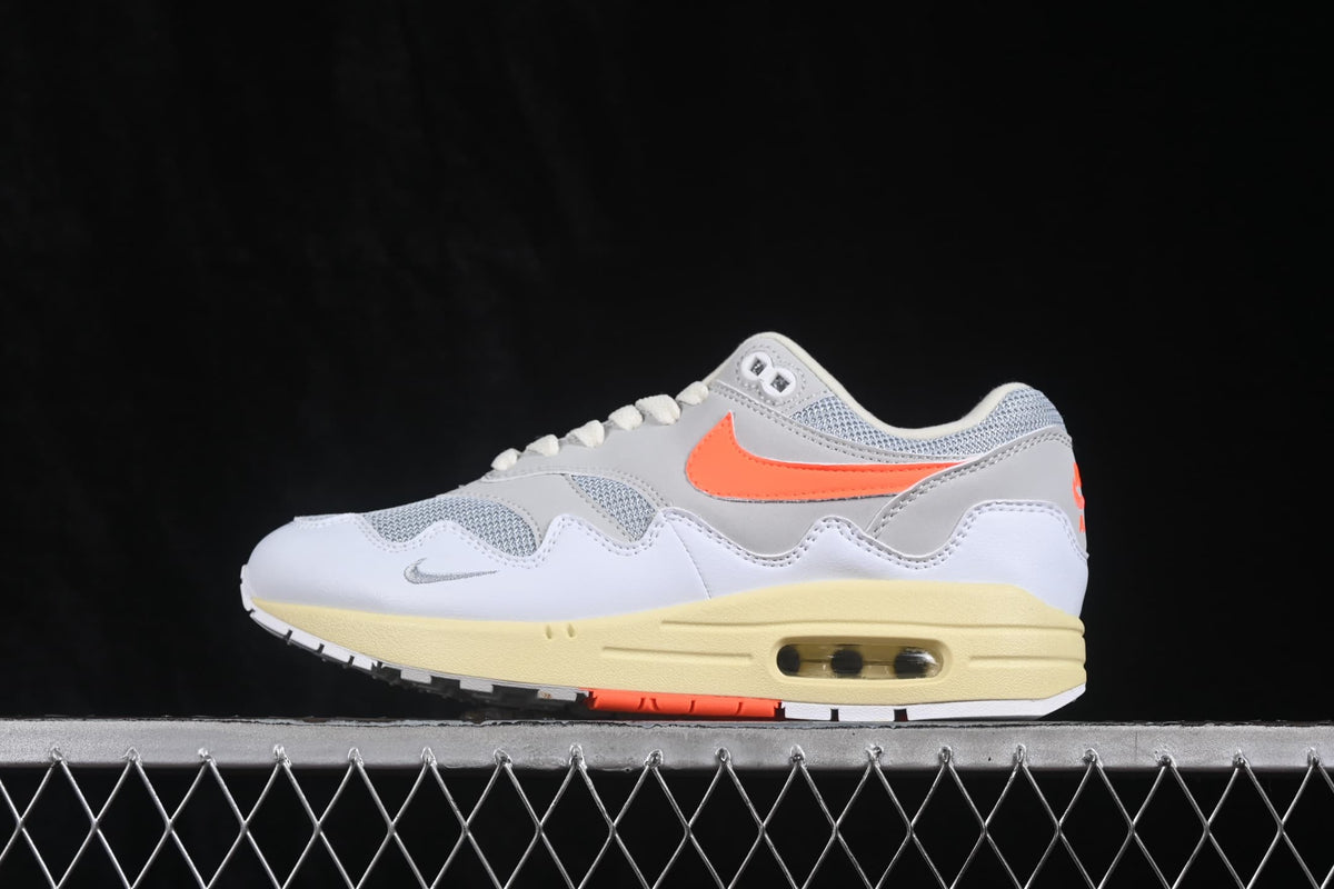 Side profile of a Nike Air Max 1 sneaker showing the orange Swoosh, grey overlays, cream-colored midsole and visible Air unit.